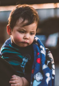 A dark haired toddler with a grumpy look holding a blanket